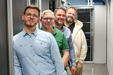 Vier männliche Personen im Serverraum die hintereinander stehen und lächelnd in die Kamera schauen